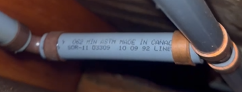 What Happens During Poly-B Replacement? A Homeowner’s Guide 1 Close-up of grey Poly-B plumbing pipes with metal crimp fittings between floor joists.